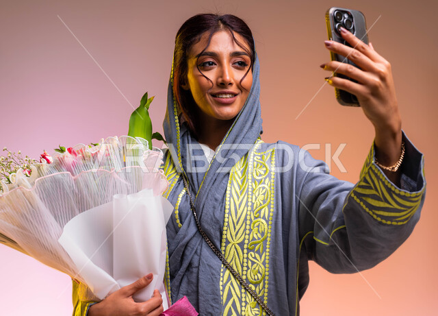إنتقاء باقات الزهور في الأعياد والمناسبات السعيدة، بورتريه لامرأة عربية خليجية سعودية ترتدي عباءة مطرزة عصرية تقوم بإلتقاط صور سيلفي تذكارية مع باقة الورود الفاخرة، إيماءات السعادة والفرح، محلات ومتاجر بيع وشراء الأزهار الطبيعية، خلفية ملونة