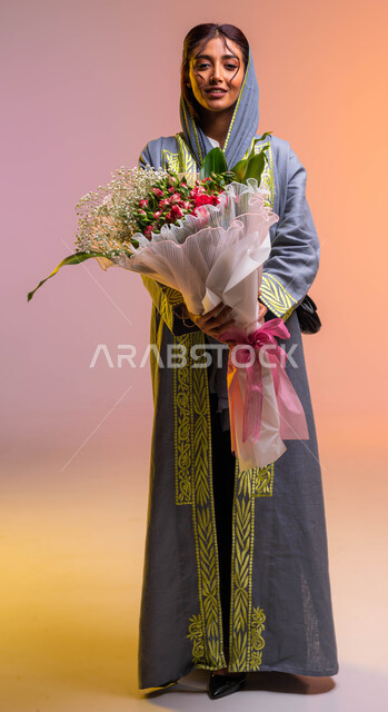 صورة الجسم بالطول الكامل, محلات ومتاجر بيع وشراء الزهور الطبيعية, إمتلاك باقات الزهور في الأعياد والمناسبات السعيدة، إيماءات تدل على الفرح والسعادة، بورتريه لإمرأة عربية خليجية سعودية ترتدي عباءة مطرزة عصرية تحمل بيدها باقة ورود فاخرة، خلفية ملونة