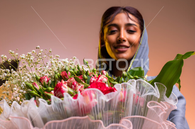 إيماءات تدل على الجمال و الأنوثة، شراء باقات الزهور في الأعياد والمناسبات السعيدة، بورتريه مقرب لشابة عربية خليجية سعودية ترتدي عباءة مطرزة عصرية تحمل بيدها باقة ورود فاخرة، محلات ومتاجر بيع وشراء الزهور الطبيعية, خلفية ملونة