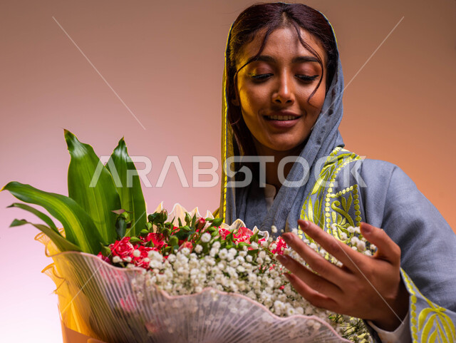إيماءات وجه ويدين مختلفة تدل على الفرح والسعادة، شراء باقات الزهور في الأعياد والمناسبات السعيدة، بورتريه مقرب لشابة عربية خليجية سعودية ترتدي عباءة مطرزة عصرية تحمل بيدها باقة ورود فاخرة، محلات ومتاجر بيع وشراء الزهور الطبيعية, خلفية ملونة
