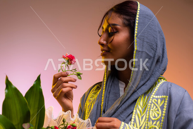 إيماءات الإعجاب بالروائح العطرية الزكية للورد، محلات ومتاجر بيع وشراء باقات الورود والهدايا في الأعياد والمناسبات السعيدة، بورتريه مقرب لشابة عربية خليجية سعودية ترتدي العباءة العصرية تقوم بشم رائحة الزهور، خلفية ملونة