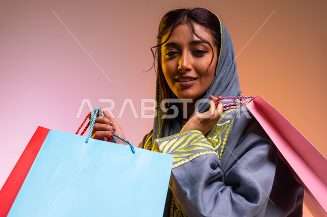 قضاء وقت ممتع في شراء المستلزمات، ايماءات السعادة والشعور بمتعة التسوق، بورتريه مقرب لإمرأة عربيه خليجية سعوديه ترتدي العباءه العصرية تحمل بيدها أكياس التسوق الملونه، الاستفادة من العروض والتخفيضات الموسميه، خلفية ملونة 