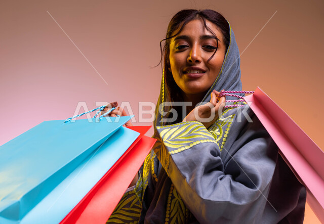 Taking advantage of seasonal offers and discounts, gestures of happiness and feeling the pleasure of shopping, a close-up portrait of a Saudi Arabian Gulf woman wearing a modern abaya carrying colored shopping bags in her hand, buying gifts and supplies, 