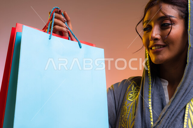 ايماءات السعادة والشعور بمتعة التسوق، الاستفادة من العروض والتخفيضات الموسميه، بورتريه مقرب لإمرأة عربيه خليجية سعوديه ترتدي العباءه العصرية تحمل بيدها أكياس التسوق الملونه, شراء الهدايا والمستلزمات، خلفية ملونة 