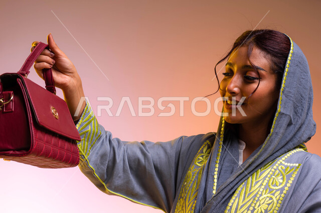 حقائب عصرية وموديلات مختلفة، منتجات نسائيه سعوديه وماركات عالمية فاخرة، متعة التسوق وشراء الحقائب الجديدة، بورتريه مقرب لإمرأة عربية خليجية سعودية ترتدي العباءة العصرية، ايماءات الإعجاب والسعادة لامتلاك حقيبة أنيقة، خلفية ملونة