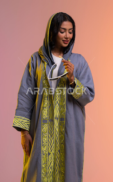 إيماءات الأنوثة والجمال، عرض تصاميم العباءات السعوديه الحديثة, الاهتمام بالموضة والأناقة، بورتريه لإمرأة عربيه سعوديه خليجيه مبتسمة تمسك النظارة الشمسية، ارتداء عباءة ملونة عصريه، شراء أحدث منتجات الملابس والاكسسوارات النسائية، خلفية ملونة