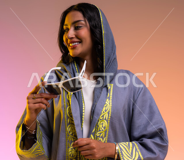 ايماءات السعادة والفرح, عرض تصاميم موديلات عباءات سعودية معاصرة, مفهوم الأناقة والاهتمام بالمظهر الخارجي, بورتريه مقرب لشابة عربية خليجية سعودية مبتسمة تمسك النظارة الشمسية, شراء الملابس والاكسسوارات الجديدة, خلفية ملونة