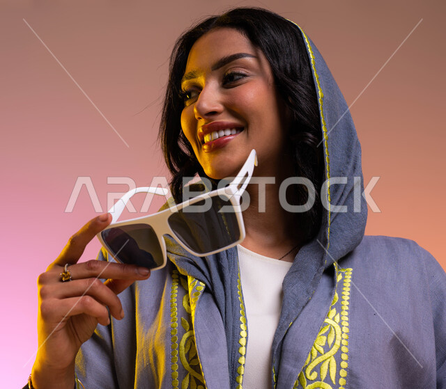مفهوم الأناقة والاهتمام بالمظهر الخارجي, ايماءات السعادة والفرح, بورتريه مقرب لشابة عربية خليجية سعودية مبتسمة تمسك النظارة الشمسية, شراء الملابس والاكسسوارات الجديدة, عرض تصاميم موديلات عباءات نسائية سعوديه معاصرة, خلفية ملونة