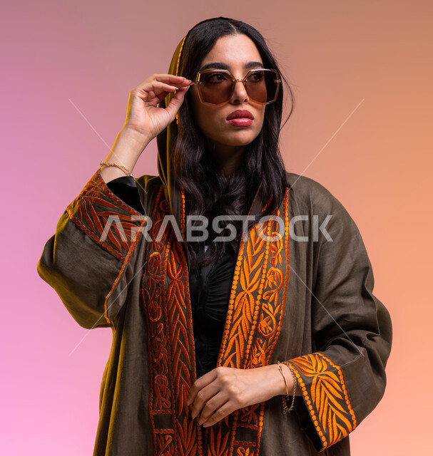 ملابس واكسسوارات فاخرة, متابعة آخر صيحات الموضة والجمال, تسوق وشراء أحدث منتجات النظارات العصرية الحديثة, بورتريه مقرب لشابة عربية سعودية خليجية تقوم بتجربة نظارة شمسية جديدة, ارتداء عباءة خليجية بنية مطرزة, الاحتراف في عرض الأزياء النسائية, خلفية ملونة