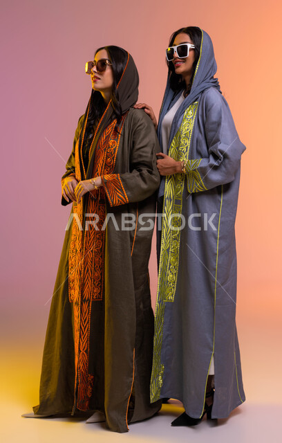 الاحتراف في عرض الأزياء, متابعة أحدث صيحات الموضة بارتداء عباءات عصرية ملونة مع نظارات شمسية عالمية, بورتريه لرفيقتان مبتسمتان عربيتان خليجيتان سعوديتان تنظران إلى شيء ما, صورة الجسم بالطول الكامل, خلفية ملونة
