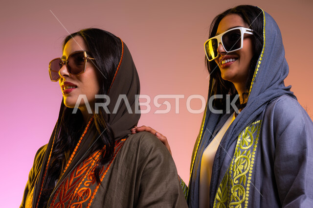 متابعة أحدث صيحات الموضة والأزياء بارتداء عباءات عصرية ملونة مع نظارات شمسية عالمية, بورتريه لصديقتان مبتسمتان عربيتان خليجيتان سعوديتان تنظران إلى شيء ما، مفهوم الموضة والأناقة، شراء الملابس والاكسسوارات الجديدة، خلفية ملونة