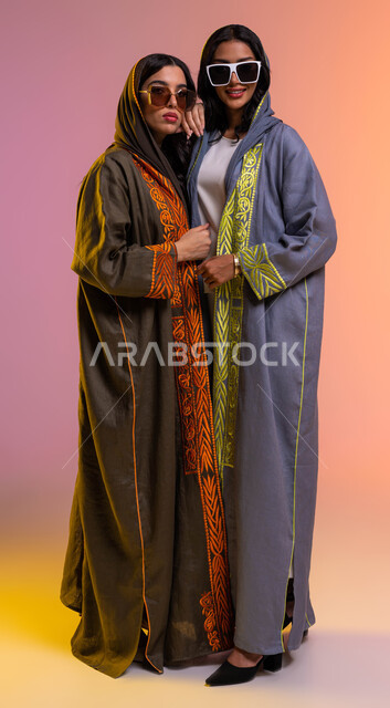 ايماءات الاحتضان والمحبة, قضاء أوقات ممتعة مع الأصدقاء، بورتريه  لصديقتان عربيتان خليجيتان سعوديتان ترتديان عباءات خليجية أنيقة وملونة ونظارات شمسية عصرية، مفهوم الموضة والأناقة، أحدث الأزياء والاكسسوارات المعاصرة، صورة الجسم بالطول الكامل, خلفية ملونة