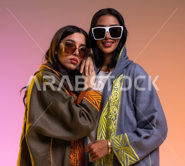 قضاء أوقات ممتعة مع الأصدقاء، ايماءات الاحتضان والمحبة, بورتريه  لصديقتان عربيتان خليجيتان سعوديتان ترتديان العباءات الخليجية العصرية والنظارات الشمسية، مفهوم الموضة والأناقة، أحدث المنتجات النسائية السعودية، خلفية ملونة