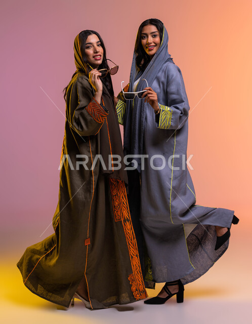 The concept of elegance and attention to the outward appearance, happy female gestures, a portrait of two Saudi Arabian Gulf female friends wearing modern Gulf abayas and elegant sunglasses, a full-length body image, Saudi women's products, a colored back