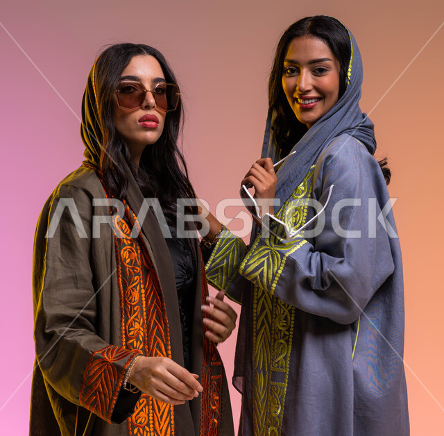 عرض الأزياء والموديلات الحديثة, ايماءات الصداقة والمحبة, ارتداء نظارات شمسية عصرية وعباءات خليجية نسائية أنيقة، بورتريه لرفيقتان عربيتان خليجيتان سعوديتان، مفهوم الموضة والأناقة، شراء الملابس والاكسسوارات الجديدة، خلفية ملونة