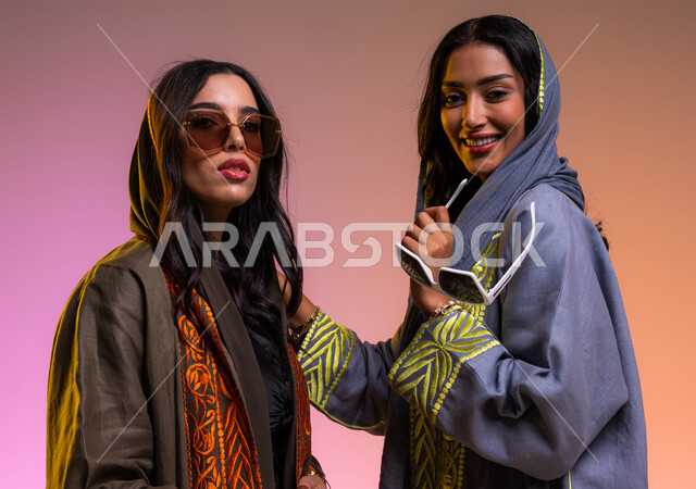 Saudi women's products, gestures of femininity and softness, wearing modern sunglasses and elegant Gulf women's abayas, a portrait of two Saudi Arab Gulf women, the concept of fashion and elegance, buying new clothes and accessories, a colored background