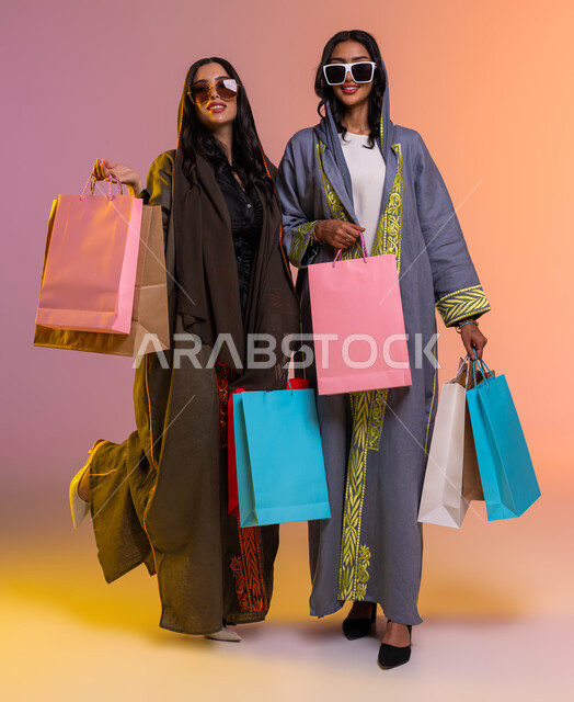 شراء أحدث منتجات النسائية من المولات التجارية، الإهتمام بالموضة والجمال والأناقة، بورتريه لصديقتان عربيتان سعوديتان خليجيتان تحملان اكياس التسوق الملونة، إرتداء العباءات الأنيقة والنظارات الشمسيه العصرية، صوره للجسم بالطول الكامل, خلفية ملونة