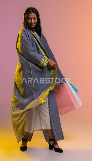 إيماءات الاعجاب بالعباءه الجديدة العصريه، صورة الجسم بالطول الكامل،  بورتريه لإمرأه عربيه سعوديه خليجيه تحمل اكياس التسوق، متابعة أحدث صيحات الموضه و الجمال، التسوق وشراء أحدث منتجات الملابس والإكسسوارات النسائية، خلفية ملونة