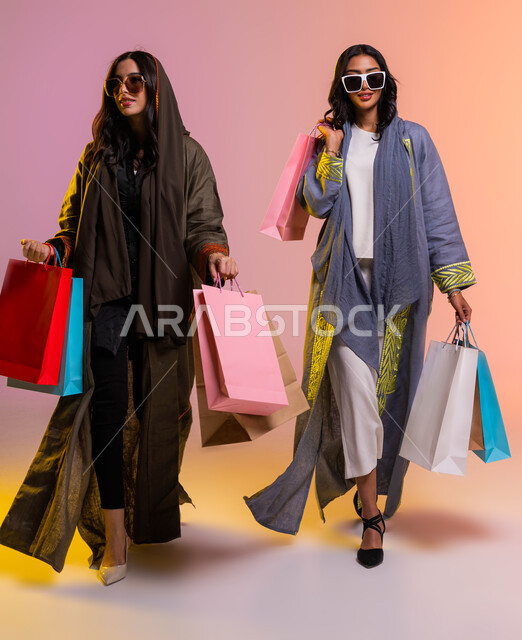 العودة من السوق التجاري، صورة للجسم بالطول الكامل،  عروض وتخفيضات موسميه، بورتريه لصديقتان عربيتان سعوديتان خليجيتان تحملان اكياس التسوق، ارتداء عباءة ملونة عصريه، التسوق وشراء أحدث منتجات الملابس والإكسسوارات النسائية، خلفية ملونة