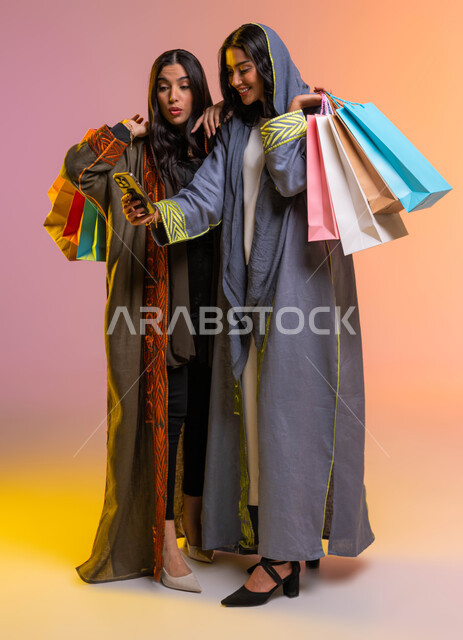 إستخدام الجوال في التسوق الإلكتروني، متعة التسوق مع الاصدقاء، الإهتمام بالموضة والجمال والأناقة، بورتريه لصديقتان عربيتان سعوديتان خليجيتان تحملان اكياس التسوق، ارتداء عباءة ملونة عصريه، التسوق وشراء أحدث منتجات الملابس والإكسسوارات النسائية، خلفية ملونة