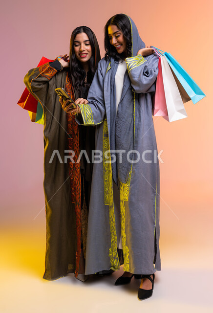  التسوق الإلكتروني عبر الهاتف المحمول، متعه التسوق مع الاصدقاء، الإهتمام بالجمال والأناقة، بورتريه لصديقتان عربيتان سعوديتان خليجيتان تحملان اكياس التسوق،  ارتداء عباءة ملونة عصريه،  خلفية ملونة، صورة الجسم بالطول الكامل