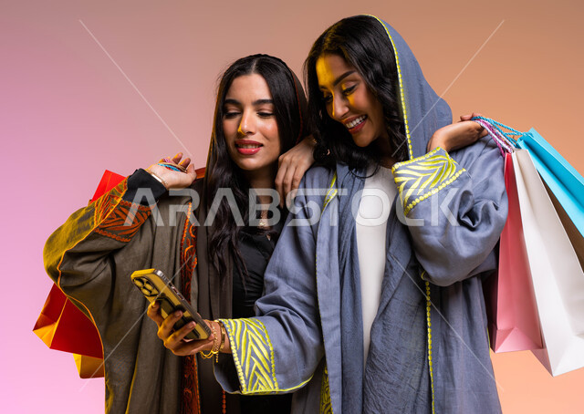 E-shopping via a mobile phone, the pleasure of shopping with friends, interest in fashion, beauty and elegance, a portrait of two Saudi Arabian Gulf female friends carrying shopping bags, wearing a modern colorful abaya, shopping and buying the latest wom