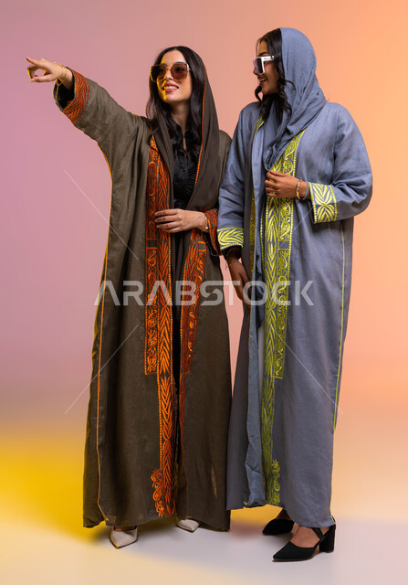صوره الجسم بالطول الكامل، عباءات خليجيه نسائيه عصريه، بورتريه لرفيقتان عربيتان خليجيتان سعوديتان، مفهوم الموضه والأناقه،  ارتداء النظرات الشمسيه،  خلفيه ملونة دردشه وحوارات بين الصديقات