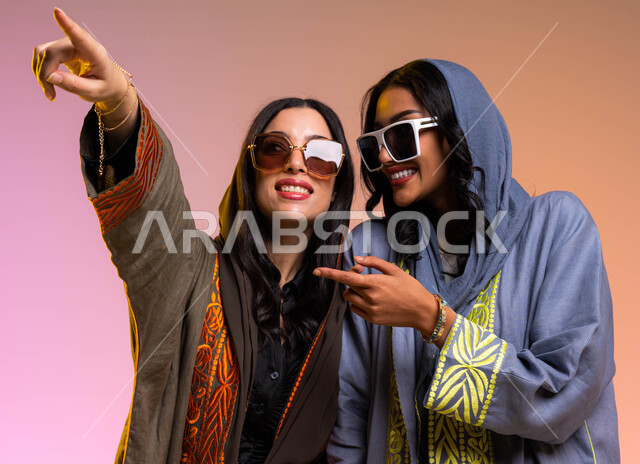 إيماءات النظر و الإشاره، عباءات خليجيه نسائيه عصريه، بورتريه لرفيقتان عربيتان خليجيتان سعوديتان، مفهوم الموضه والأناقه، ارتداء النظرات الشمسيه، خلفيه ملونة، دردشه ومحادثات بين الصديقات