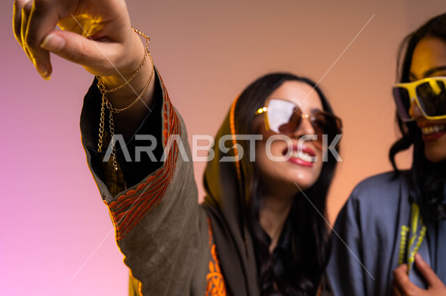 إيماءات الإشاره الى شيئ ما، عباءات خليجيه نسائيه عصريه، بورتريه لرفيقتان عربيتان خليجيتان سعوديتان، مفهوم الموضه والأناقه، ارتداء النظرات الشمسيه، خلفيه ملونة، دردشه و محادثات بين الصديقات