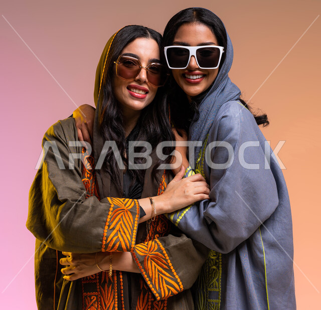 مفهوم الموضه والأناقه، إيماءات الصداقه الحميمه و الحب و المودة،  إرتداء النظارات الشمسيه و العباءات الخليجيه و الإكسسورات النسائيه العصريه، بورتريه مقرب لرفيقتان عربيتان خليجيتان سعوديتان تحتضنان،  خلفيه ملونة
