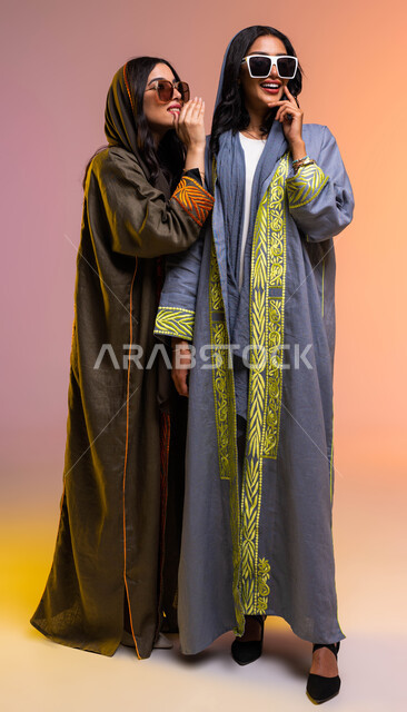 كتمان وحفظ الأسرار، التحدث بصوت منخفض، بورتريه لسيدتان عربيتان خليجيتان سعوديتان، ارتداء العباءة الخليجية والنظارات الشمسية، سيدة تهمس بأذن صديقتها، المحافظة على سرية العمل، ايماءات وجه ويدين تدل على السعادة، صورة الجسم بالطول الكامل، خلفية ملونة