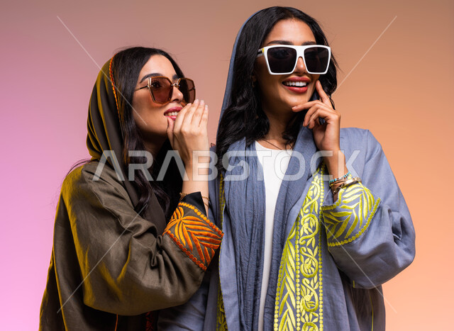 المحافظة على سرية العمل، كتمان وحفظ الأسرار، التحدث بصوت منخفض، بورتريه لسيدتان عربيتان خليجيتان سعوديتان، ارتداء العباءة الخليجية والنظارات الشمسية، سيدة تهمس بأذن صديقتها، ايماءات وجه ويدين تدل على السعادة، خلفية ملونة