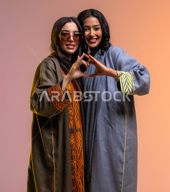 المشاركة الايجابية والعلاقات اجتماعية، ايماءات يدين عل شكل قلب، بورتريه لسيدتان عربيتان خليجيتان سعوديتان مبتسمتان، تعارف وصداقات مبنية على الحب والمودة، المحبة والترابط بين الأصدقاء، ارتداء العباءة الخليجية والنظارات الشمسية، خلفية ملونة