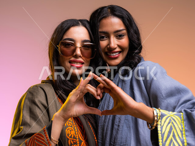 ايماءات يدين عل شكل قلب، بورتريه لسيدتان عربيتان خليجيتان سعوديتان مبتسمتان، المشاركة الايجابية والعلاقات اجتماعية، تعارف وصداقات مبنية على الحب والمودة، المحبة والترابط بين الأصدقاء، ارتداء العباءة الخليجية والنظارات الشمسية، خلفية ملونة
