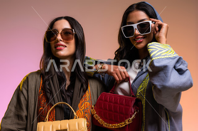 Gestures indicating femininity and beauty, buying modern bags and different models, Saudi women's products, portraits of two Saudi Arabian Gulf women, elegant Gulf gowns, wearing sunglasses, the concept of fashion and elegance, a colored background