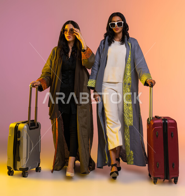 عرض موديلات جديدة لحقائب السفر والملابس والاكسسوارات، بورتريه لإمرأتان عربيتان سعوديتان خليجيتان ترتديان عباءات خليجية نسائية عصرية، مفهوم السياحة والسفر، الاستعداد لقضاء عطلة سعيدة مع الأصدقاء، صورة الجسم بالطول الكامل، خلفية ملونة