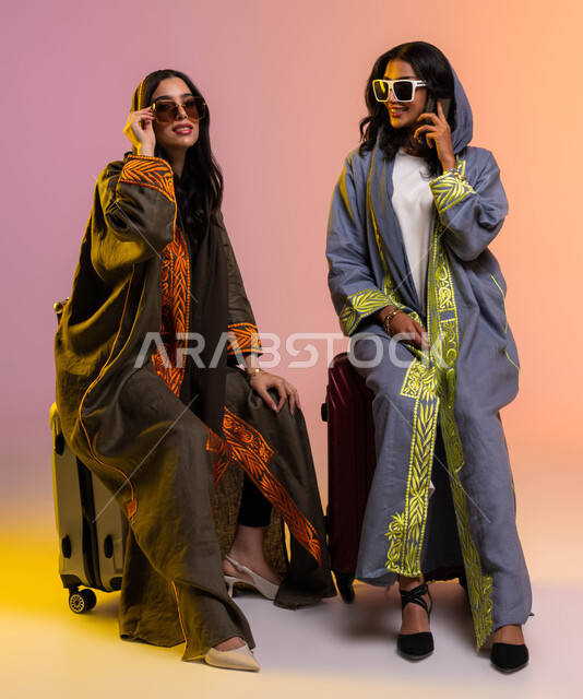 إجراء مكالمة هاتفية بواسطة الهاتف المحمول، بورتريه لرفيقتان عربيتان خليجيتان سعوديتان ترتديان عباءات خليجية نسائية عصرية، شراء حقائب السفر والملابس والاكسسوارات، الاستعداد للسفر في العطلة الصيفية، صورة الجسم بالطول الكامل، خلفية ملونة