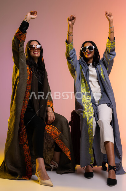 ايماءات تدل على السعادة والفرح والحماس، السفر مع الأصدقاء في العطلة الصيفية، بورتريه لرفيقتان عربيتان خليجيتان سعوديتان ترتديان عباءات خليجية نسائية عصرية، شراء حقائب السفر والملابس والاكسسوارات ، ارتداء نظارات شمسية، خلفية ملونة