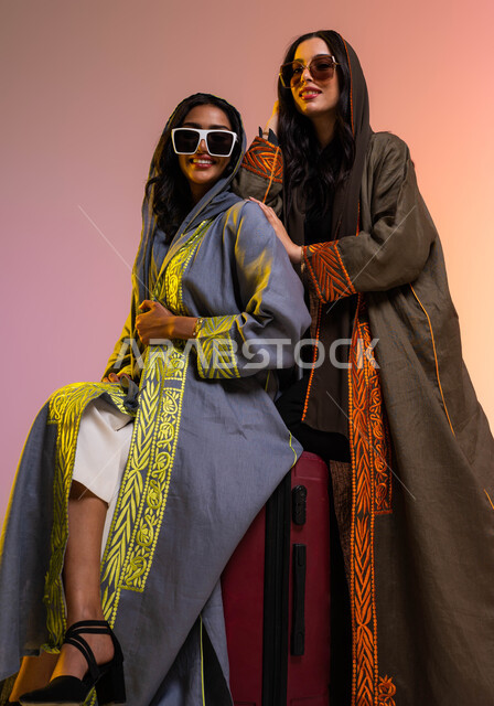 قضاء أوقات ممتعة مع الأصدقاء، ايماءات الفرح والسعاة والحماس للسفر، بورتريه لشابتان عربيتان خليجيتان سعوديتان ترتديان العباءات الخليجية العصرية و النظارات الشمسية، الجلوس على حقيبة السفر، الاستعداد لعطلة صيفية سعيدة، التجهيز للسفر، خلفية ملونة