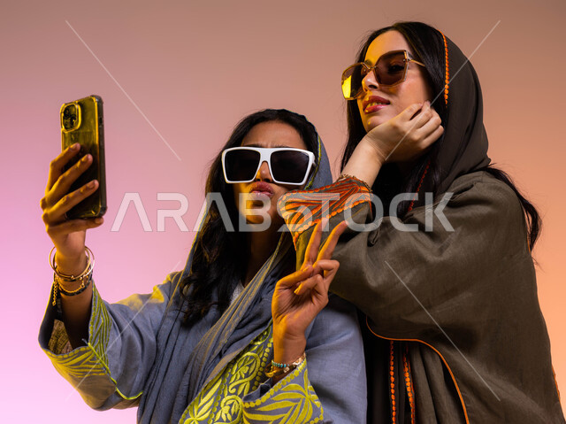 ايماءات انوثة شبابية ظريفة، مفهوم التصوير والشهرة عبر برامج التواصل الاجتماعي، التقاط صورة سلفي عبر الجوال، بورتريه لرفيقتان عربيتان خليجيتان سعوديتان ترتديان عباءات خليجية عصرية ونظارات شمسية، قضاء أوقات ممتعة مع الأصدقاء، خلفية ملونة