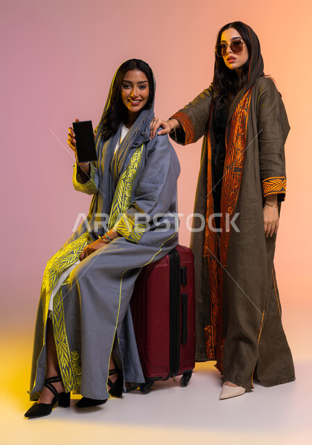 ايماءات السعادة بالتسوق الآمن عبر الاونلاين، عرض شاشة سوداء باستخدام الهاتف المحمول، بورتريه لرفيقتان عربيتان خليجيتان سعوديتان ترتديان العباءة الخليجية، الاستعداد والتجهيز للسفر، عطلة صيفية ممتعة، صورة الجسم بالطول الكامل, خلفية ملونة