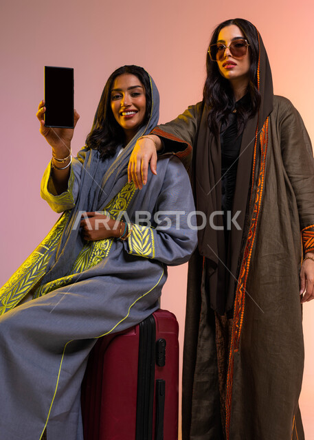 الحجز الالكتروني للسفر والسياحة عبر الهاتف المحمول، عرض شاشة سوداء فارغة على الجوال، عطلة صيفية ممتعة مع الأصدقاء، بورتريه لصديقتان عربيتان خليجيتان سعوديتان ترتديان عباءات واكسسوارات نسائية عصرية، خلفية ملونة