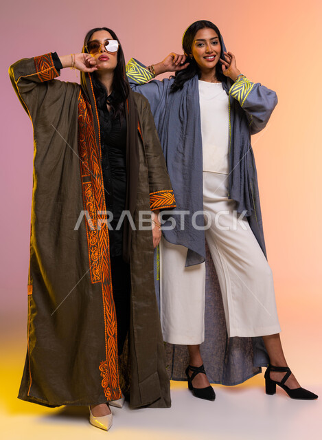 عباءات خليجية نسائية عصرية، بورتريه لعارضتان عربيتان خليجيتان سعوديتان، مفهوم الموضة والأناقة، عرض الأزياء والموديلات الحديثة، ارتداء النظارات الشمسية، شراء الملابس والاكسسوارات الجديدة، صورة الجسم بالطول الكامل، خلفية ملونة