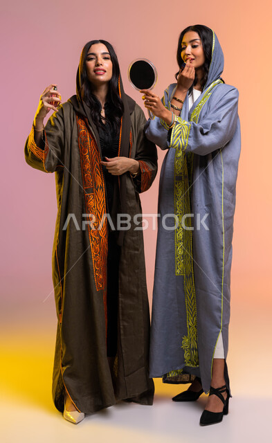 التجمل بقلم أحمر الشفاه من المستحضرات النسائيه العالميه، بورتريه لصديقتان عربيتان سعوديتان خليجيتان تستمتعان بالتزيين النسائي، مفهوم الأناقة والإهتمام بالمظهر الخارجي، التطيب والتعطر بالعود والزيوت العطرية، صورة الجسم بالطول الكامل, خلفية ملونة