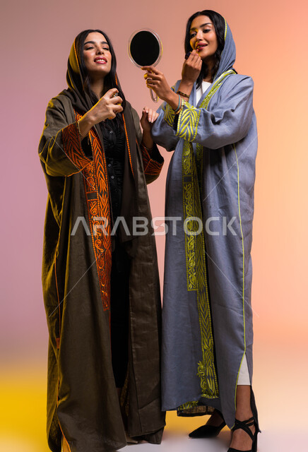 استخدام قلم أحمر الشفاه من مستحضرات نسائيه عالميه، بورتريه لصديقتان عربيتان سعوديتان خليجيتان تستمتعان بالتزيين النسائي، مفهوم الأناقة والإهتمام بالمظهر الخارجي، التطيب والتعطر بالعود والزيوت العطرية، صورة الجسم بالطول الكامل, خلفية ملونة