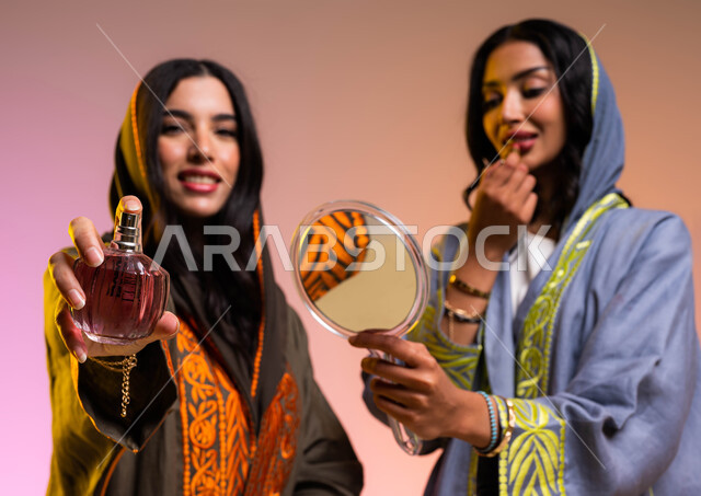 التطيب والتعطر بالعود والزيوت العطريه، استخدام مستحضرات نسائيه عالميه، مفهوم الأناقة والإهتمام بالمظهر الخارجي، بورتريه لإمرأتان عربيتان سعوديتان خليجيتان تستمتعان بالتزيين النسائي، التجمل بوضع أحمر الشفاه, خلفية ملونة