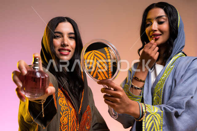 مفهوم الأناقة والإهتمام بالمظهر الخارجي، ايماءات الاستمتاع بالتزيين النسائي، بورتريه لإمرأتان عربيتان سعوديتان خليجيتان تقومان بوضع مستحضرات تجميل نسائيه عالميه، التطيب والتعطر بالعود والزيوت العطرية، استخدام قلم أحمر الشفاه, خلفية ملونة