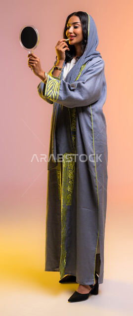 صورة الجسم بالطول الكامل، خلفية ملونة لمرأة خبيرة بمستحضرات التجميل، التسويق لمنتجات ومستحضرات المكياج في المتاجر و عبر الإنترنت، مفهوم الإهتمام بالجمال والبشرة، بورتريه لخبيرة ميكآب عربية خليجية سعودية تضع قلم أحمر الشفاه عبر النظر إلى ىالمرأة