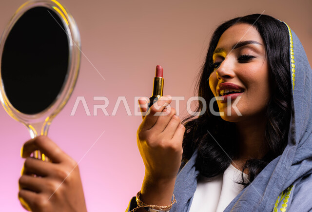 ايماءات الاستمتاع بوضع المكياج, الشغف بوضع الميكآب, أدوات مكياج تتوريال، مفهوم الإهتمام بالجمال والبشرة، التسويق لمنتجات ومستحضرات التجميل، بورتريه لخبيرة تجميل عربية خليجية سعودية تحمل إحدى أدوات الميكآب (قلم أحمر الشفاه) وتنظر إلى المرآة، خلفية ملونة