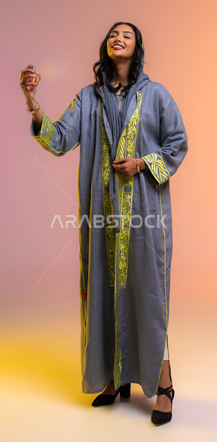 الشغف برش العطورات, مفهوم الأناقة والإهتمام بالمظهر الخارجي، بورتريه لإمرأه عربيه سعودية خليجية مبتسمة  تقوم بالتطيب والتعطر بالعود و الزيوت العطرية، التسويق لمنتجات العطور والمستحضرات النسائيه, صورة الجسم بالطول الكامل، خلفية ملونه